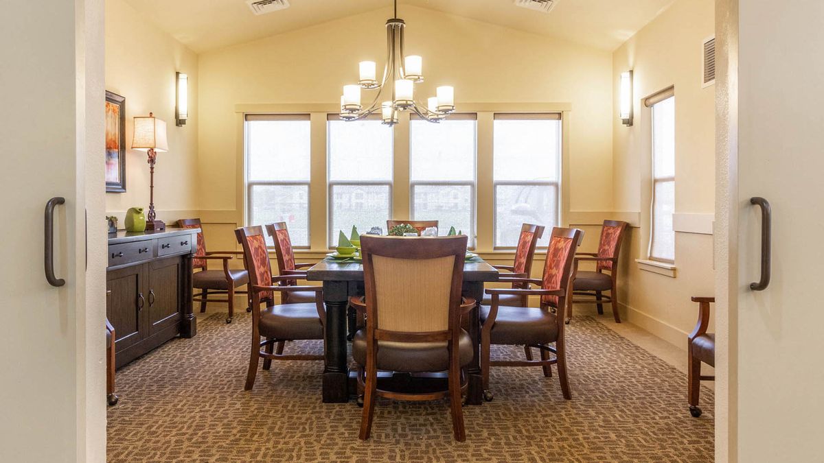 A well-lit dining room with a large table surrounded by chairs, featuring multiple windows and a central chandelier.