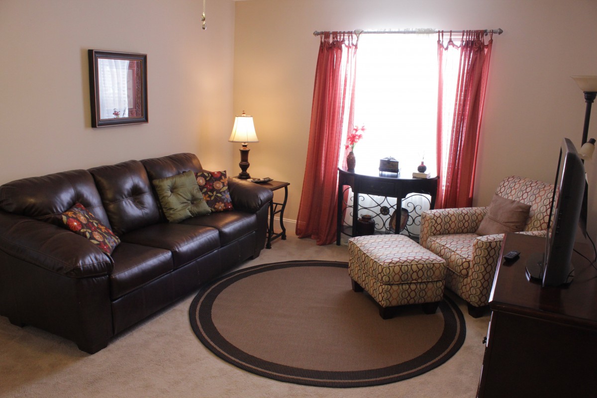 A cozy living room with a brown leather sofa, a patterned armchair and ottoman, warm lighting from a lamp, and red curtains framing the window.