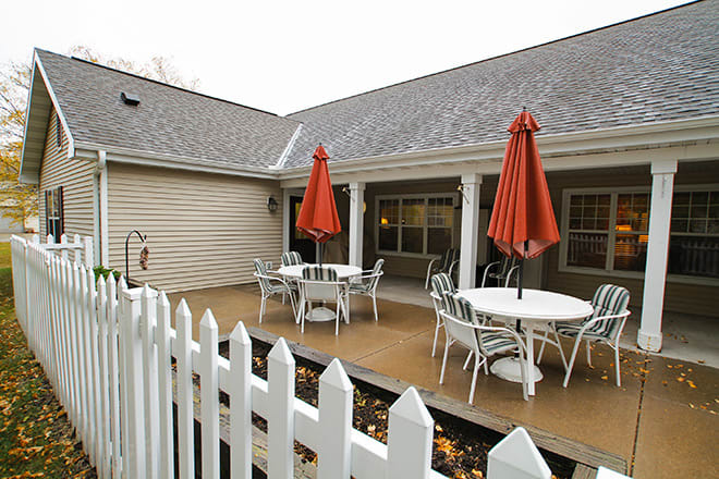 A cozy outdoor patio area with tables, chairs, and red umbrellas surrounded by a white picket fence.