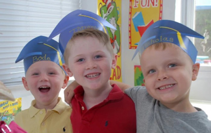 Three young children wearing graduation caps and smiling in a classroom setting.