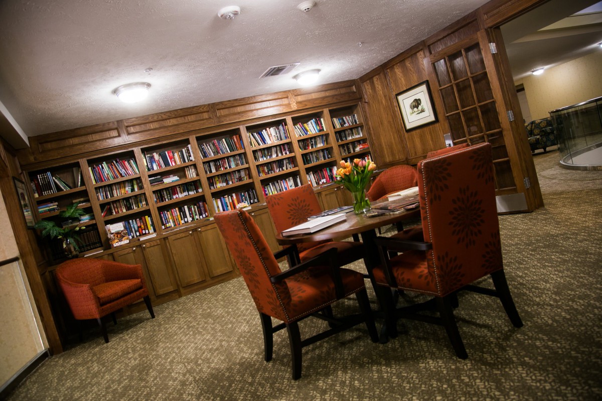 A cozy library room with bookshelves lining the walls, a table surrounded by chairs, and elegant wood paneling.