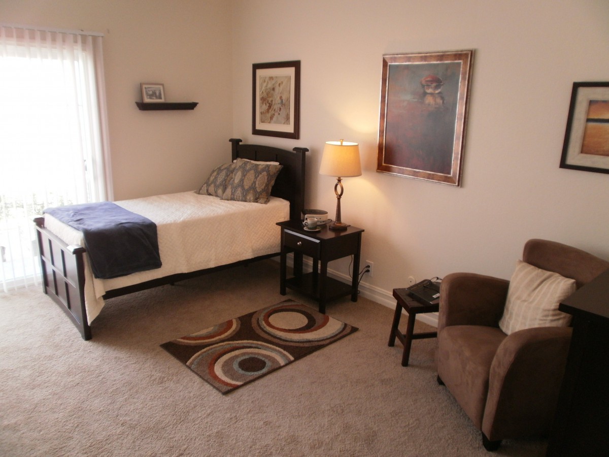 A cozy, minimalist bedroom with a single bed, lamp, and comfortable chair.