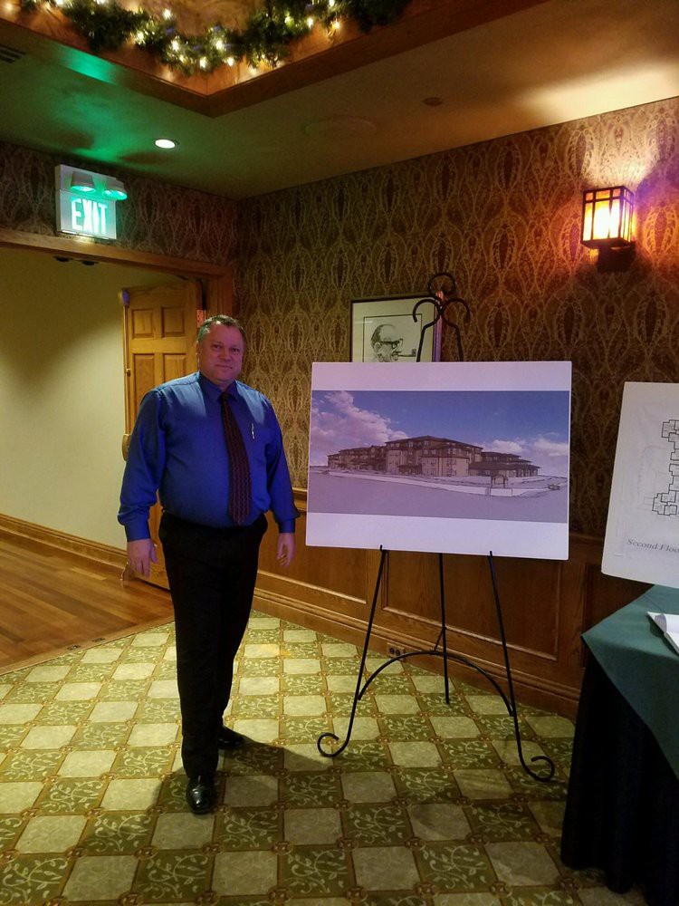 A person in formal attire stands next to an easel displaying artwork indoors.