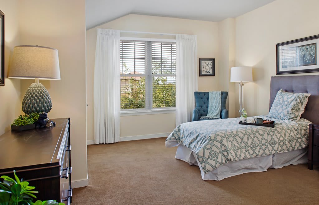 A cozy bedroom with a large window, featuring a bed with patterned bedding and a comfortable chair.