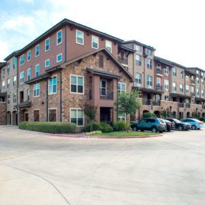 A multi-story apartment building with a parking lot and several cars parked in front.
