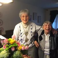 Two elderly women smiling together in a warmly lit room with a bouquet of flowers.