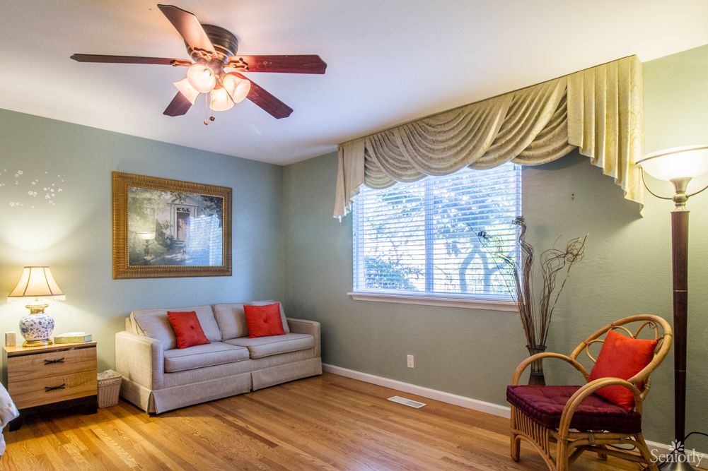 A cozy living room with a couch, an armchair, a ceiling fan, and a window with draped curtains.