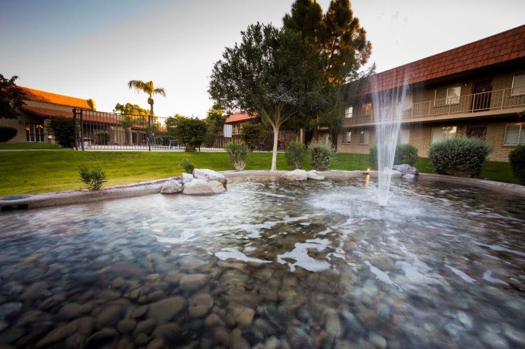 A serene outdoor landscape featuring a fountain and pond surrounded by trees and buildings.