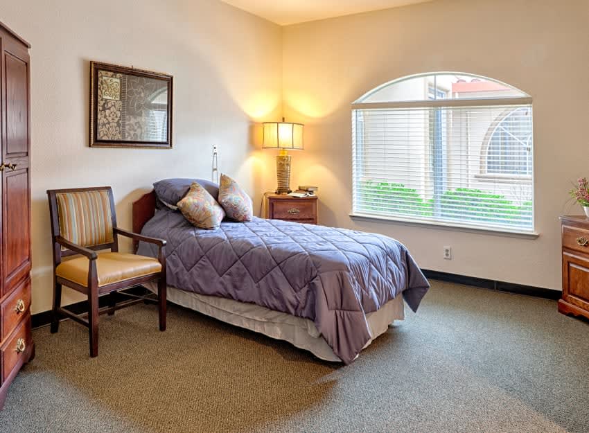 A cozy bedroom with a single bed, lamp, chair, and large window.