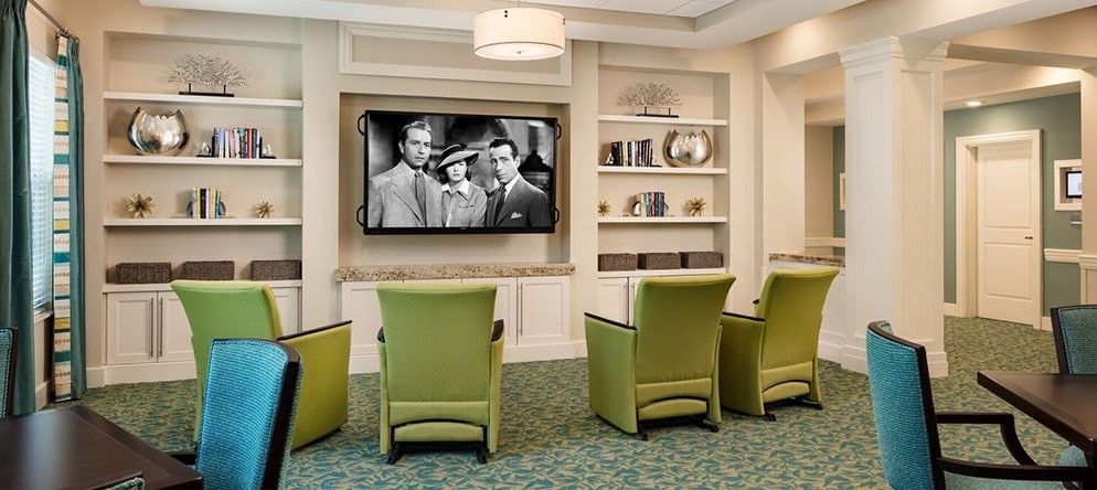 A cozy living room with green chairs facing a television displaying a black and white movie, surrounded by bookshelves.
