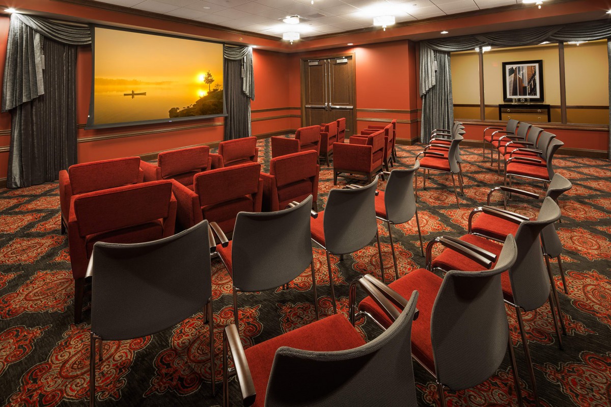 An empty conference room with red and black chairs facing a projector screen, featuring a sunset image.