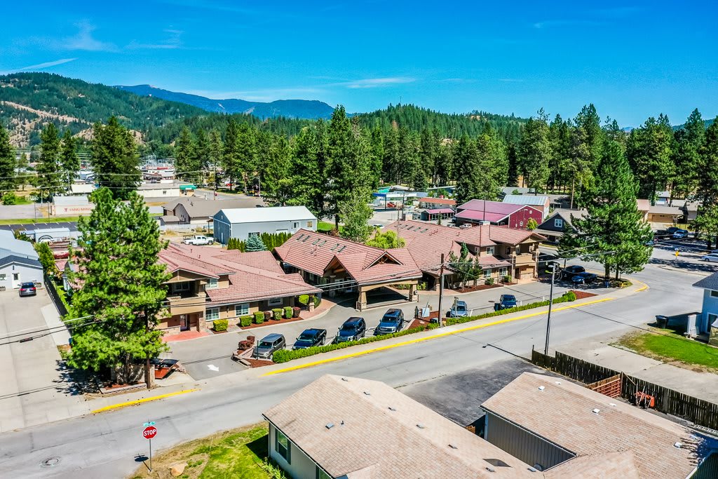 A small town with buildings and a road lined with cars, set against a backdrop of mountains and trees under a clear blue sky.