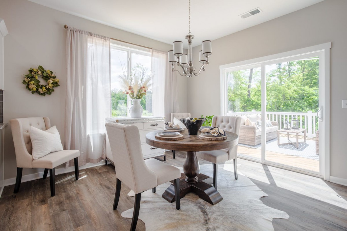 A bright and elegantly decorated dining room with a round table, upholstered chairs, and a chandelier, illuminated by natural light from large windows.