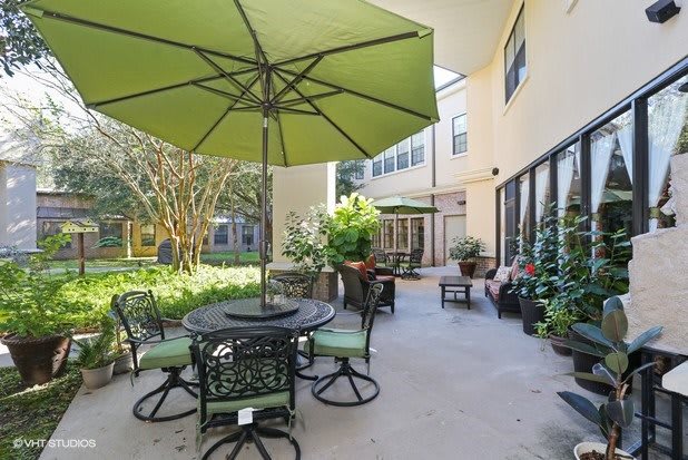 A well-furnished outdoor patio area with a table, chairs, and a large green umbrella surrounded by greenery.
