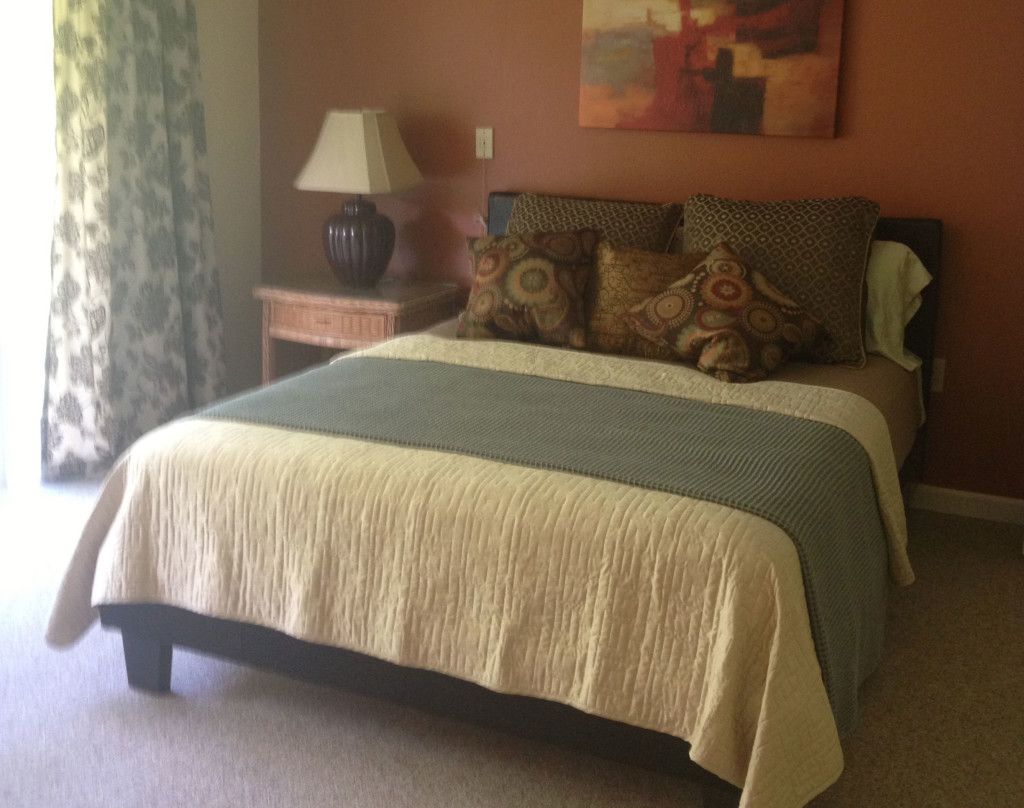 A cozy bedroom featuring a well-made bed with decorative pillows, a lamp on a wooden nightstand, and an abstract painting on the wall.