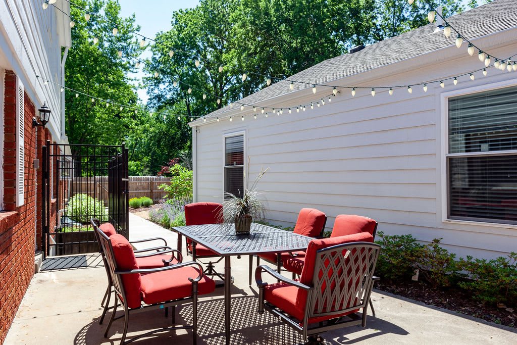 A cozy patio setup with red-cushioned chairs around a table, adorned with string lights in a sunny backyard setting.