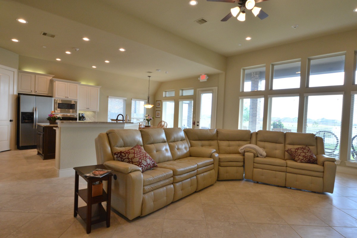 A spacious living room with a beige sofa set, open to the kitchen area and illuminated by natural light from large windows.