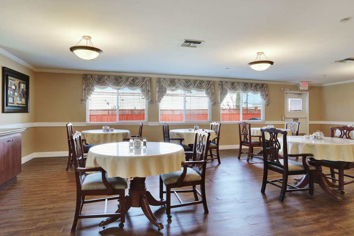 An empty dining room with tables and chairs on a wooden floor, featuring large windows with curtains.