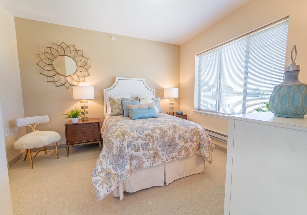 A cozy and well-decorated bedroom with neutral tones, featuring a bed with patterned bedding, two bedside lamps, and a large window letting in natural light.