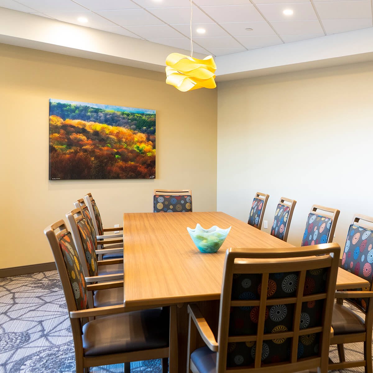 A modern conference room with a large wooden table surrounded by chairs, featuring artwork on the wall and a decorative light fixture.