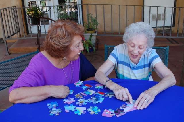 Two elderly women working on a puzzle together at an outdoor table.