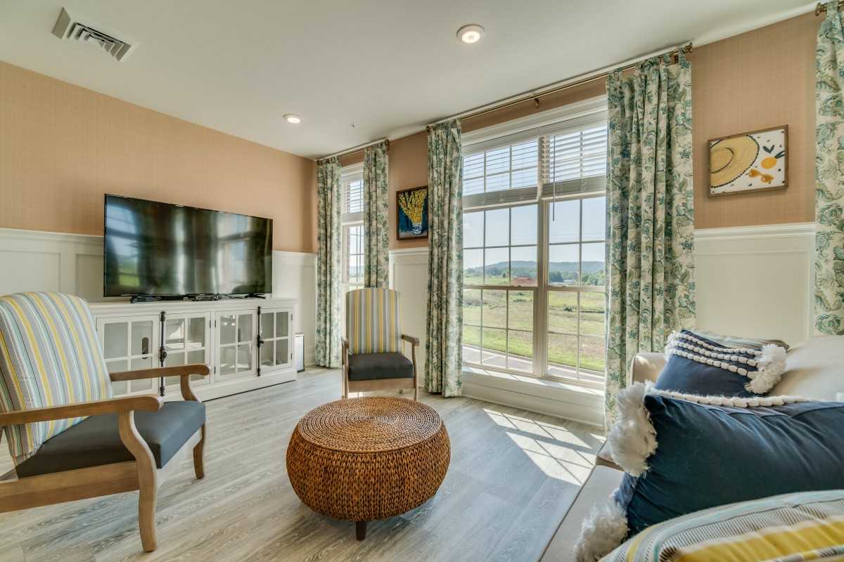 A cozy living room with a large window letting in natural light, featuring a television, comfortable seating, and a woven coffee table.