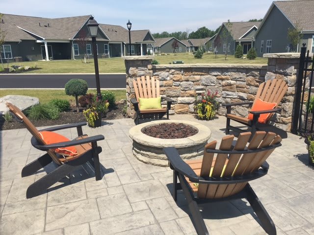 A cozy outdoor patio area with a fire pit surrounded by Adirondack chairs, set against a backdrop of landscaped gardens and charming homes.