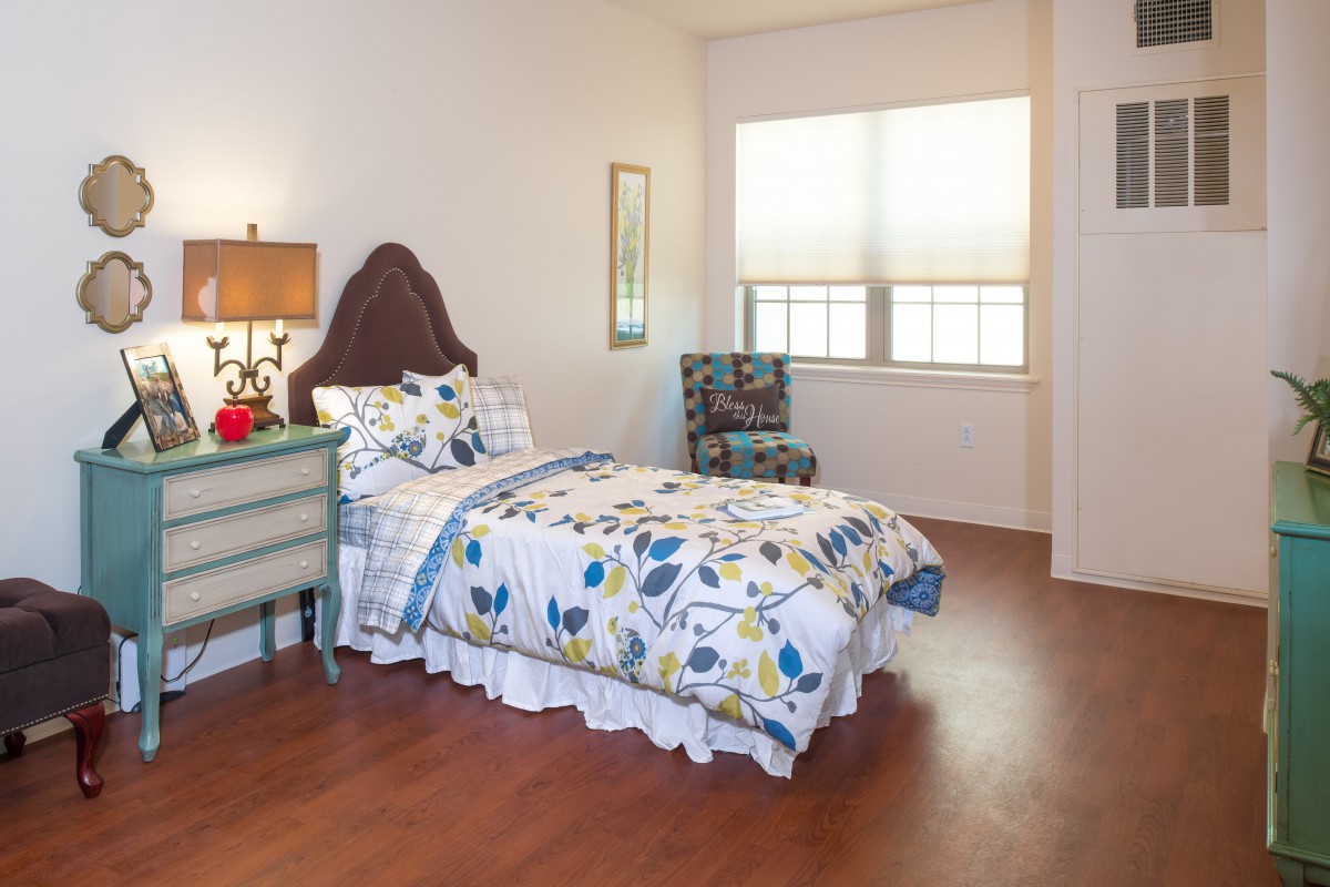 A cozy bedroom with a single bed featuring floral bedding, complemented by a nightstand and lamp, plus an armchair near the window.