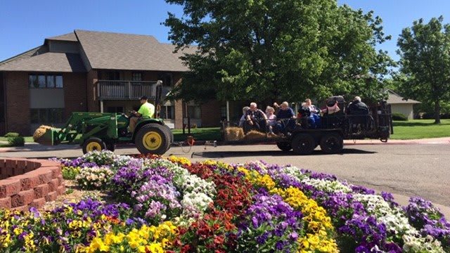 A tractor pulls a group of people on a hayride past vibrant flowerbeds in a sunny garden setting.