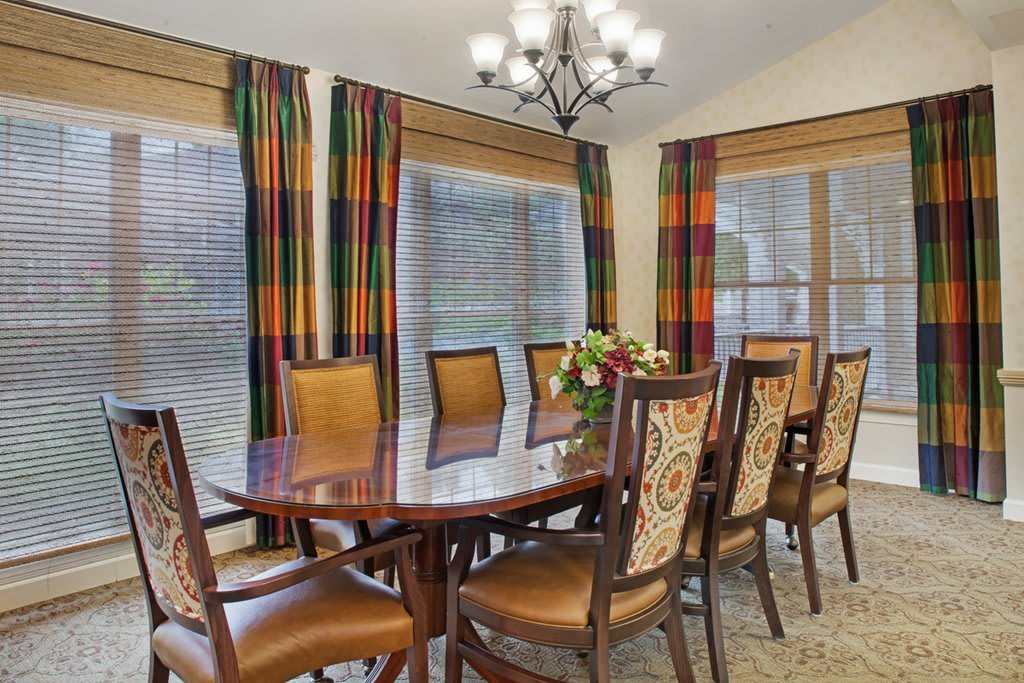A well-furnished dining room with a wooden table and chairs, accented by colorful curtains and an elegant chandelier.
