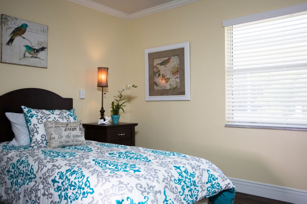 A cozy bedroom with a floral-patterned bedspread, a bedside table with a lamp and plant, and wall artwork.