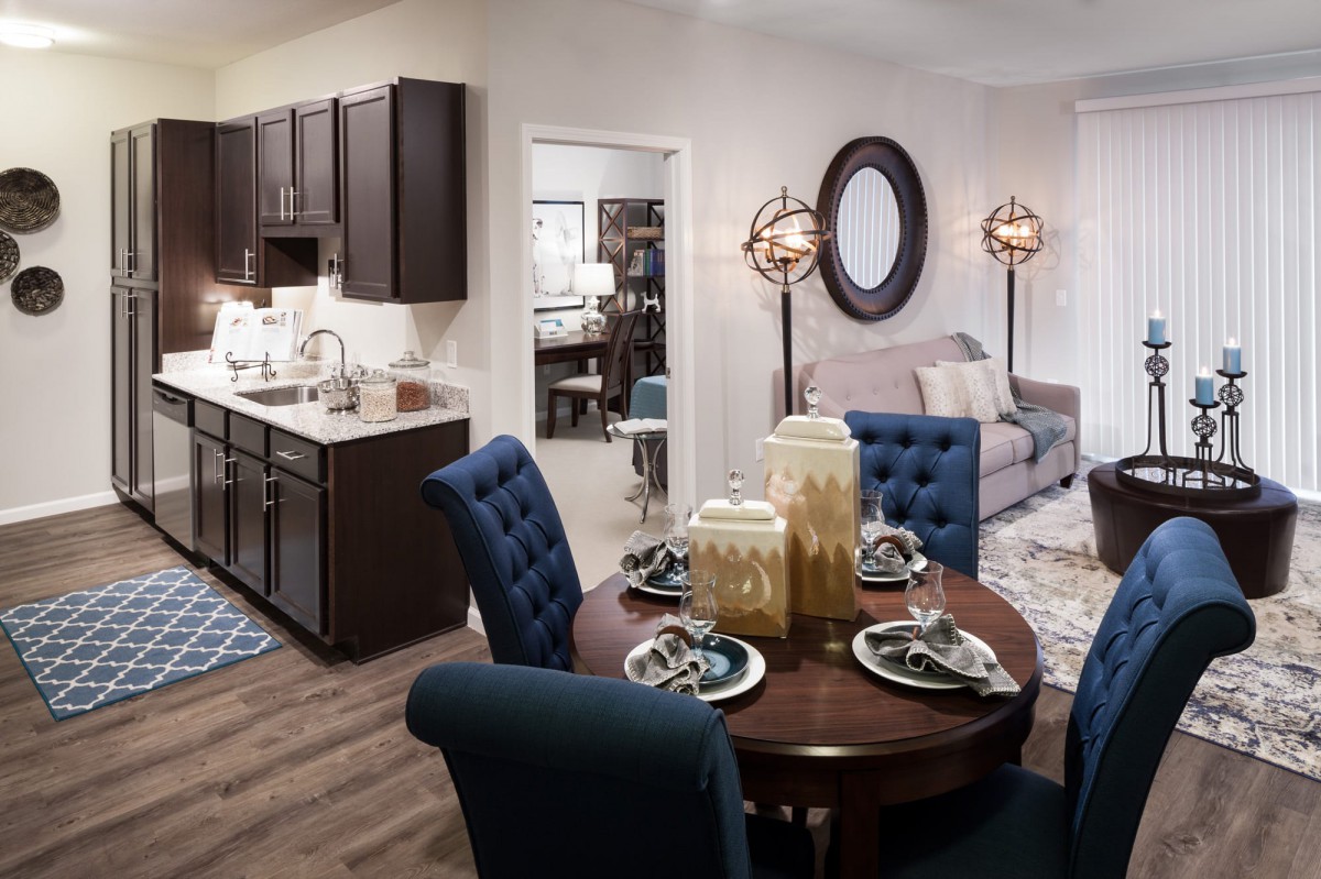 A modern apartment interior featuring a dining area with a round table and blue chairs, adjacent to a kitchen with dark cabinets and a cozy living room.