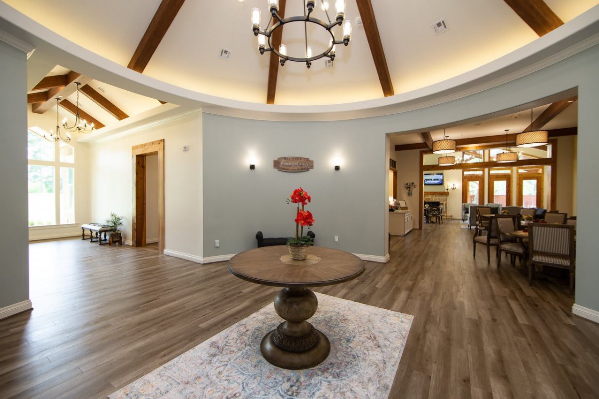 A modern lobby featuring a round wooden table, chandelier, and decorative vase under elegant wooden beams.