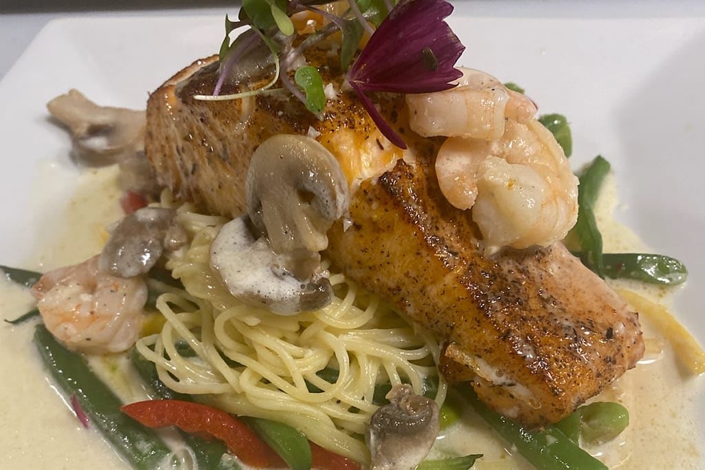 A gourmet plate featuring grilled salmon, shrimp, and mushrooms served over a bed of pasta with fresh vegetables.