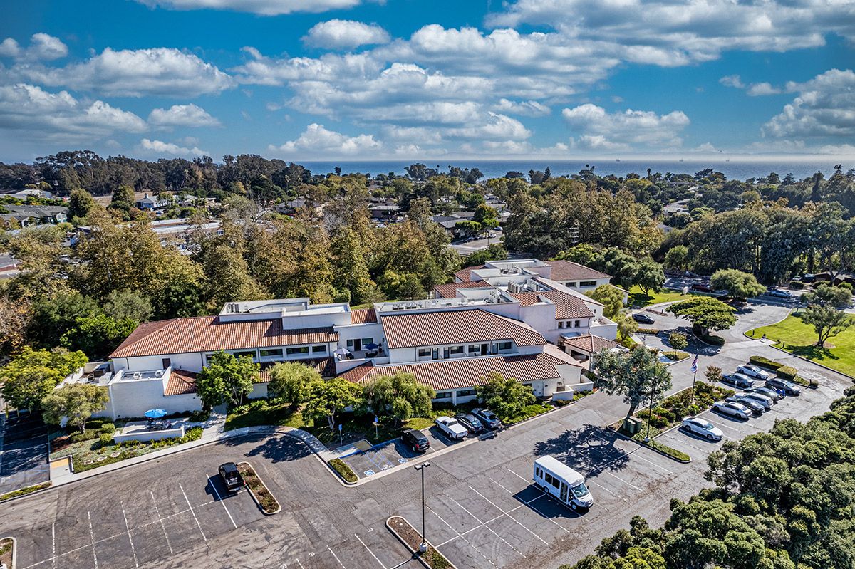 An aerial view of buildings surrounded by trees with a parking lot in the foreground under a partly cloudy sky.