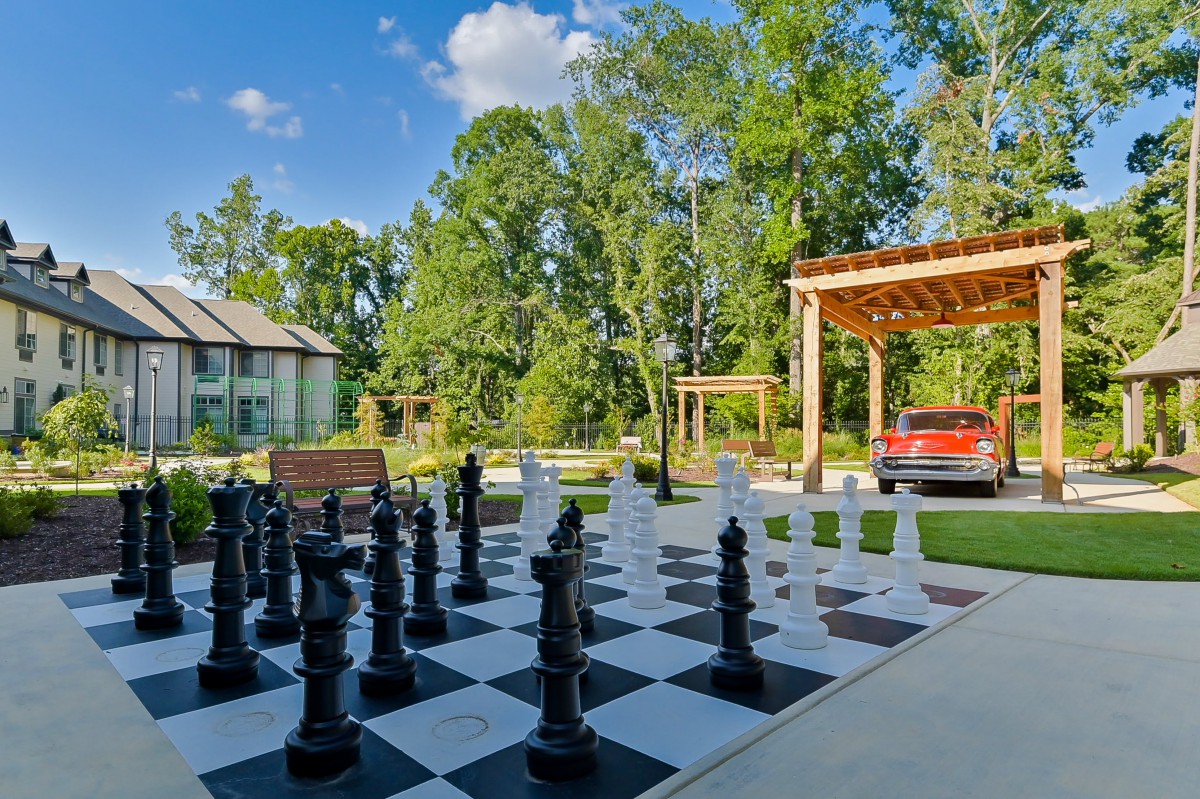 A scenic outdoor setting featuring a giant chessboard surrounded by trees, with a house and pergola in the background.