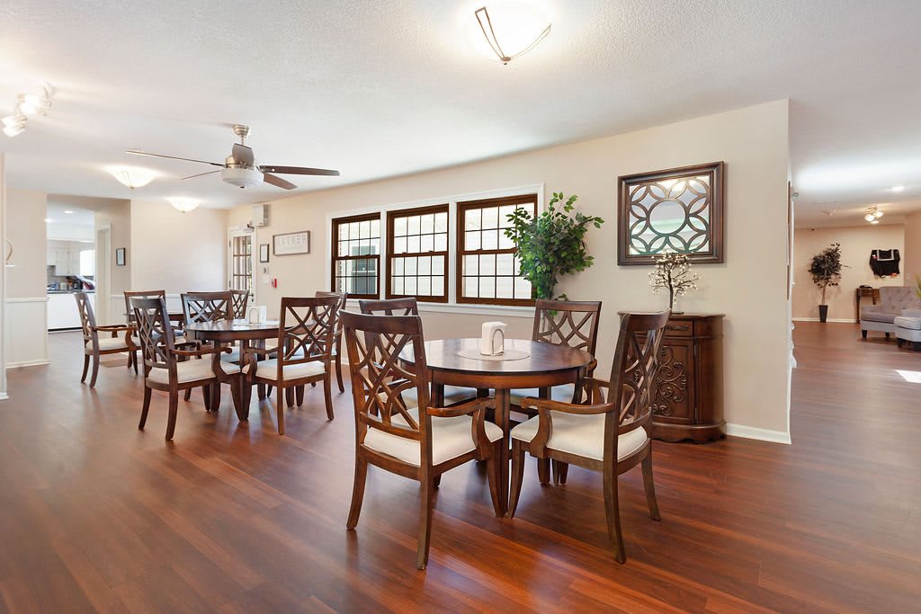 A spacious dining room with wooden furniture and flooring, featuring large windows and a decorative mirror on the wall.