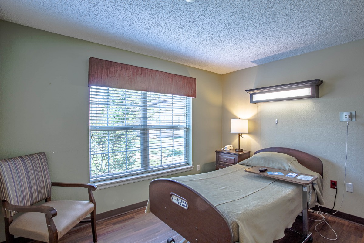 A well-lit room featuring a hospital bed, a window with curtains, a lamp, and a chair.