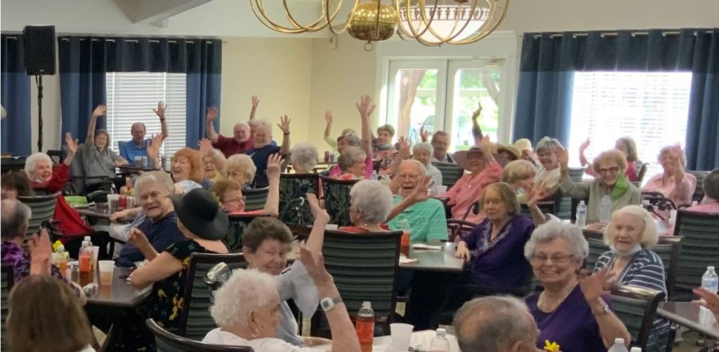 A group of seniors participating in an indoor community activity, some raising their hands enthusiastically.