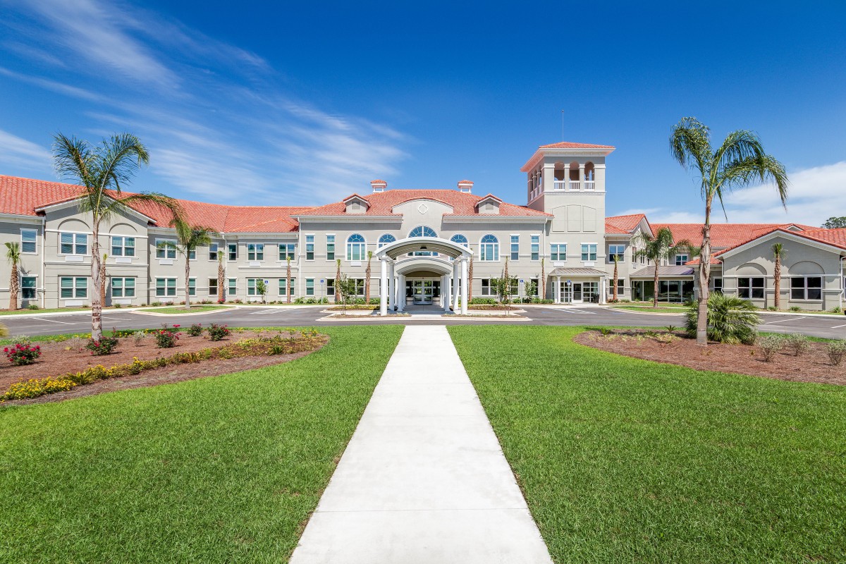 A grand building with red roof tiles and a central tower, surrounded by palm trees and a well-manicured lawn, under a clear blue sky.