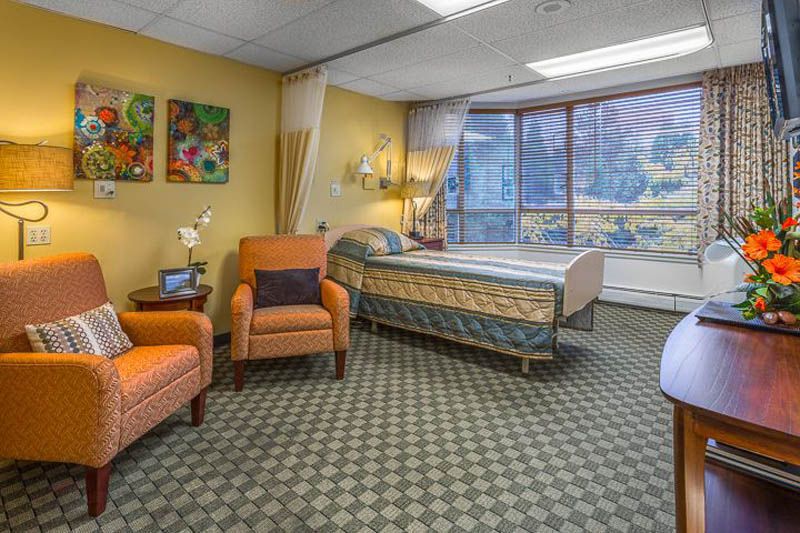 A cozy hospital room with a bed, chairs, and large windows letting in natural light.