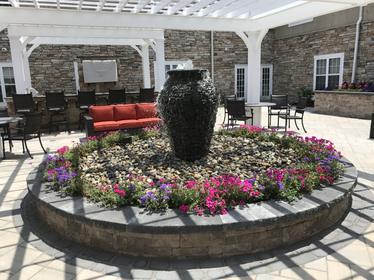 A beautiful garden patio featuring a central water fountain surrounded by vibrant flowers and comfortable seating under a pergola.