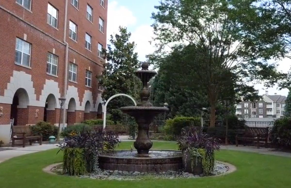 A serene garden featuring a beautiful fountain surrounded by lush greenery and a brick building.