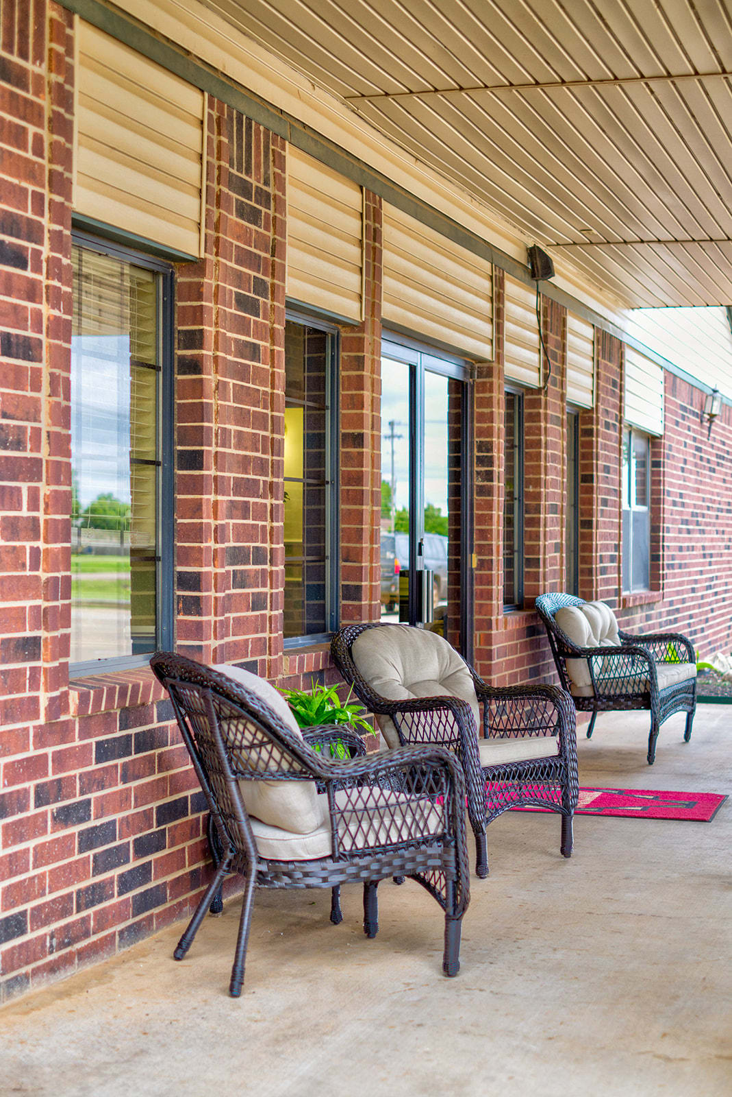 A cozy porch with wicker chairs, cushions, and a red doormat against a brick wall.