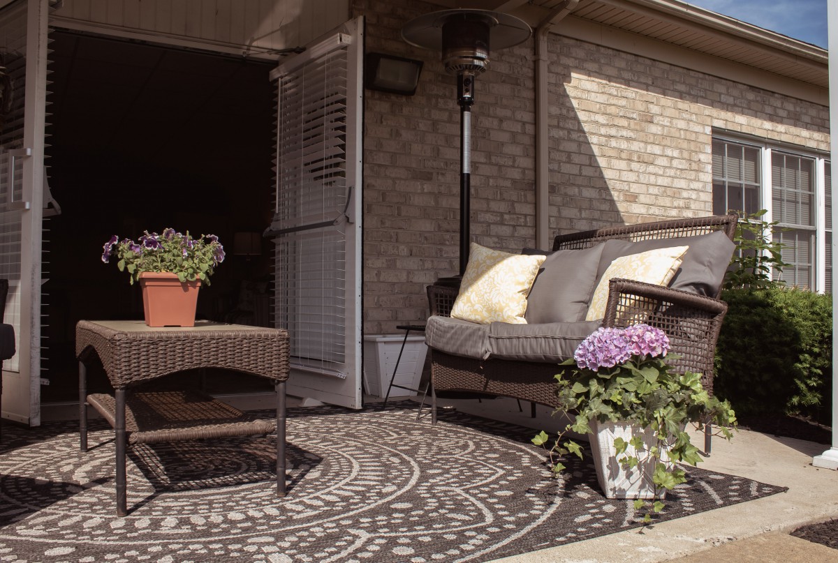 A cozy patio area with rattan furniture, potted flowers, and a patterned rug against a brick wall.