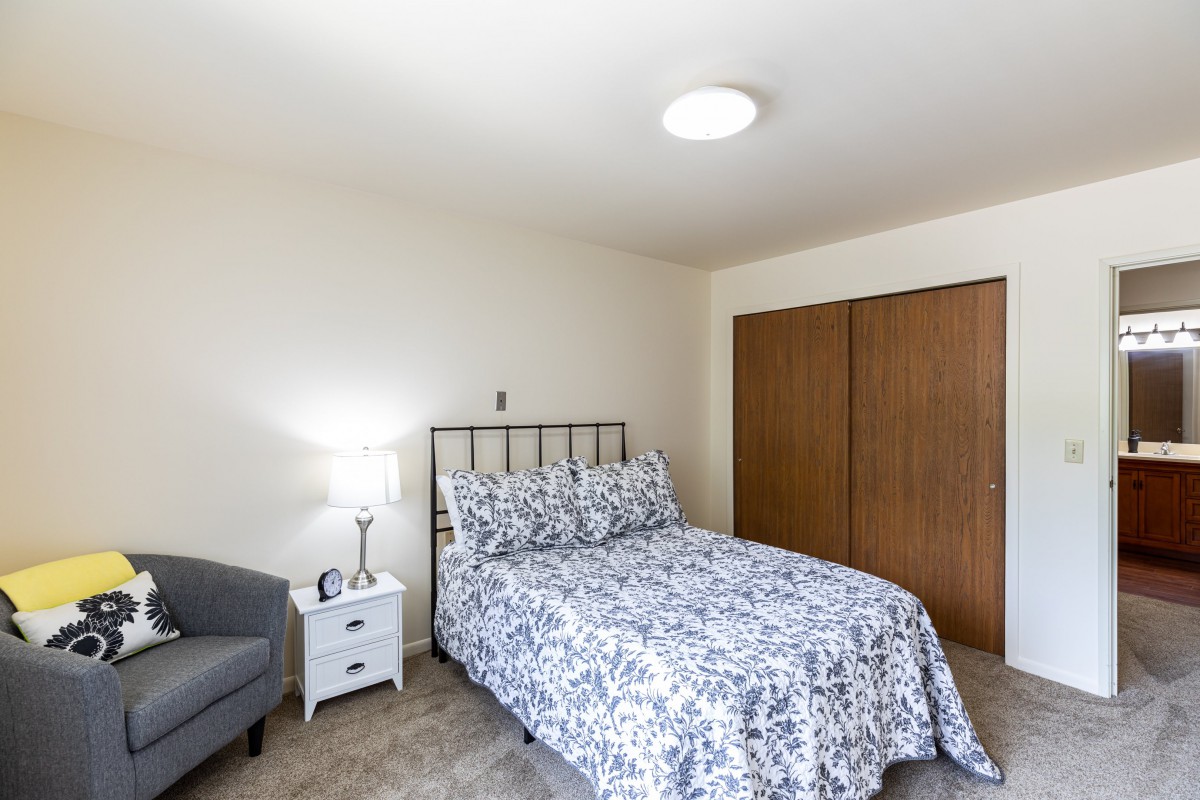 A cozy bedroom with a patterned bedspread, nightstand with lamp, chair, and wooden closet.