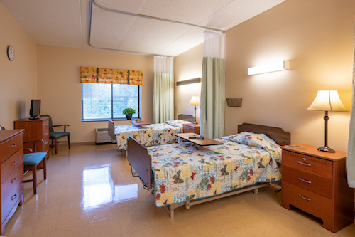 A spacious patient room in a hospital featuring two beds, warm lighting, and a view outside.
