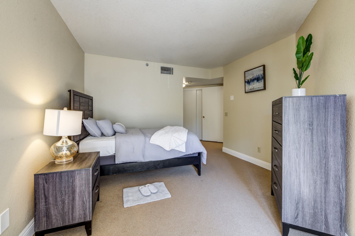 A cozy bedroom featuring a bed with pillows and a blanket, bedside table with lamp, dresser, and wall decor.