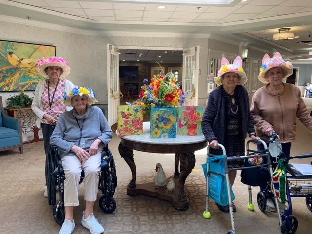 A group of elderly women wearing festive hats, with some using wheelchairs and walkers, gathered around a decorated table indoors.