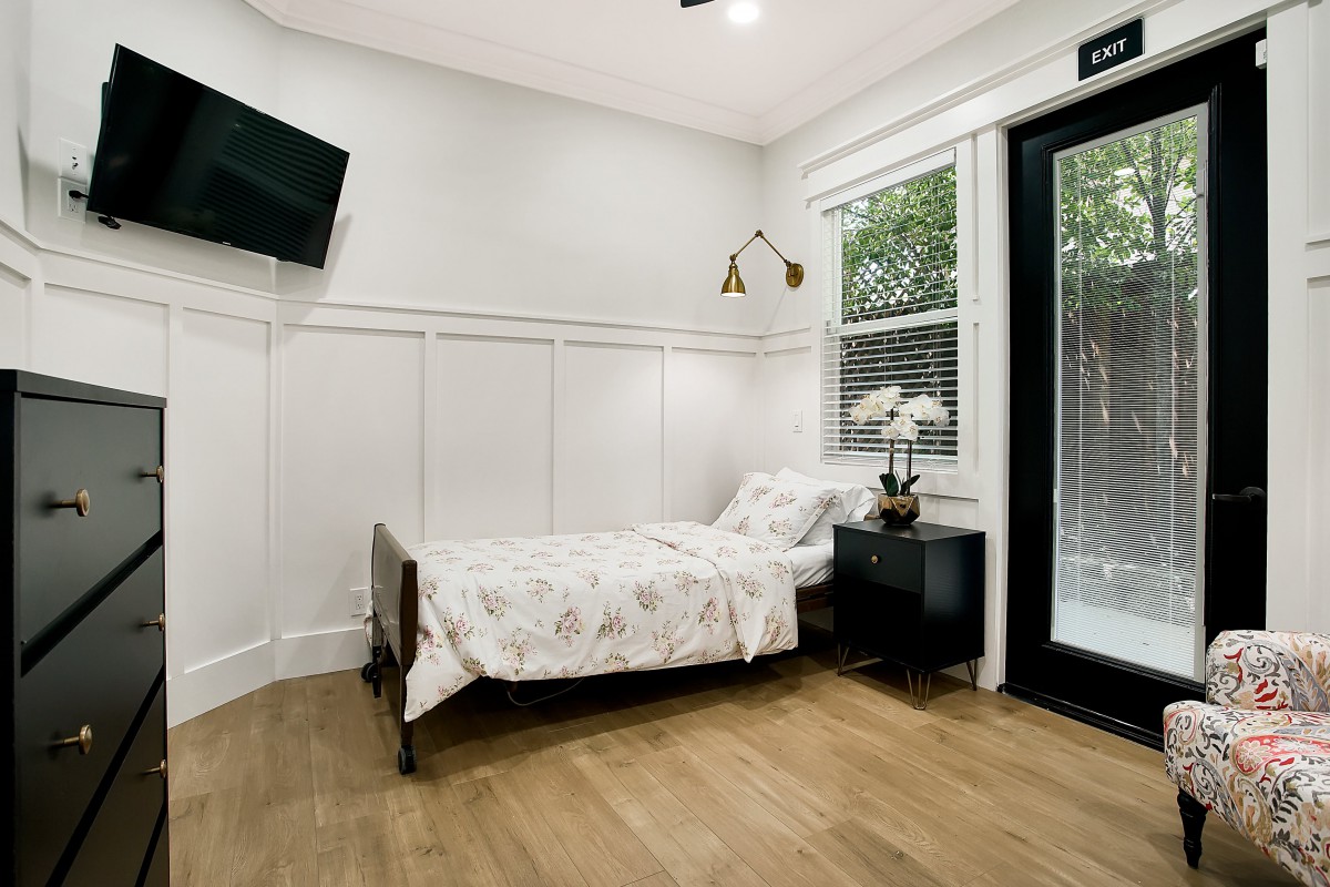 A minimalist modern bedroom with a single bed, wall lamp, and TV, featuring a wooden floor and access to natural light through a window and glass door.
